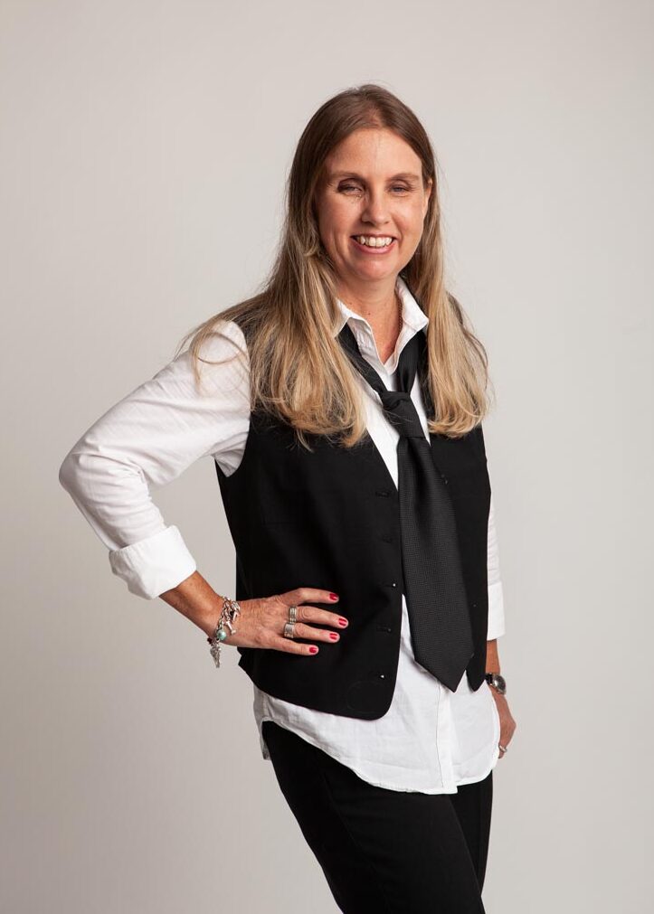 Profilbild på Anna Bergholtz. Anna står i fotostudio med långt blont hår, klädd i svart väst, vit skjorta, slips, svarta byxor och tittar mot bilden med ett leende. Fotograf: Helena Berzelius