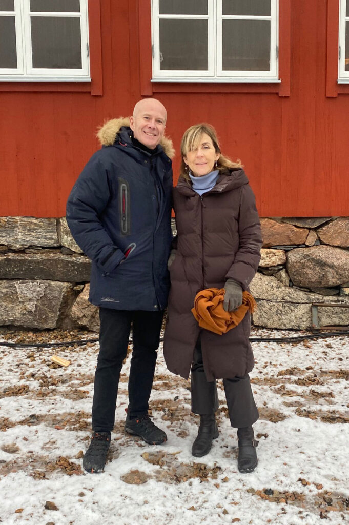 Kenth Åkerman och Andrea Hartmann Jovell står framför ett rött hus med vita fönsterramar. Marken är lätt snöklädd och Kenth och Andrea har vinterkläder på sig.