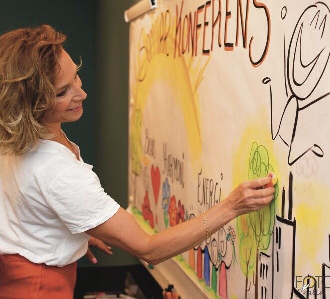Frida ritar på en färgglad whiteboard under en sommarkonferens, där hon använder en grön tuschpenna för att lägga till detaljer i en lekfull illustration av en person och byggnader. Hon ler, ser fokuserad och nöjd ut, klädd i en vit T-shirt och rostfärgade byxor.